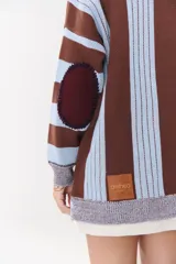 Buzo de tejido de punto con diseño de rayas verticales en tonos bordó y rojo. Presenta cuello alto con cierre frontal, mangas largas con coderas en contraste y detalles de costuras visibles.