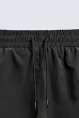 Bañador de hombre negro, largo hasta la rodilla, con acabado aterciopelado. Presenta cintura elástica ajustable con cordón, bolsillos laterales y un bolsillo trasero de plastrón. Incluye forro interior.