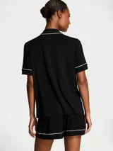 Pijama corto de modal negro compuesto por una camisa de manga corta con botones, bolsillo en el pecho y cuello solapa, y un short a juego con cintura elástica y cordón ajustable. Ambas piezas cuentan con ribetes blancos en contraste.