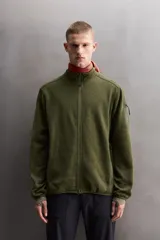 Chaqueta verde militar de punto con interior de polar. Tiene cuello alto, manga larga con bolsillo con cierre en la manga, bolsillos laterales con cierre y cierre frontal con cremallera.
