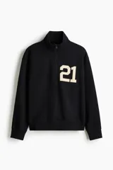 Buzo negro de tejido pesado con interior cepillado suave, cuello elevado con medio cierre y hombros ligeramente caídos. Presenta un estampado frontal con el número "21" en color beige. Puños y bajo en punto acanalado.