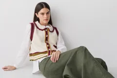 Chaleco de lana color marrón con cuello estilo bebé, cierre frontal con botones grandes y detalle de puntilla en cuello, sisas y bolsillo frontal. Presenta una franja decorativa en color rojo en el ruedo.