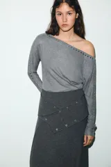 Camiseta de cuello barco y manga larga tipo murciélago, confeccionada en tejido de punto gris melange. Presenta detalle de tachas metálicas a lo largo de los hombros y las mangas.