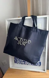 Bolso tote de tela color negro, amplio y flexible, con doble asa para el hombro y estampa frontal con la leyenda "Actitud Sur".
