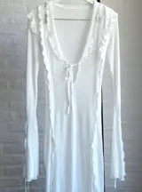 Vestido largo de microtul blanco, con escote en V profundo y mangas largas acampanadas. Presenta aberturas laterales en la falda y cierre con lazos en el frente. Es una prenda transparente.