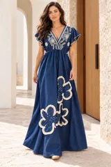 Vestido largo de denim azul con escote en V, mangas cortas con volados y cordón ajustable con borlas en la cintura. Presenta bordados detallados en el pecho y apliques grandes de flores con flecos en la parte inferior de la falda.
