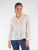 Blusa blanca de broderie de manga larga, con cuello camisero, cierre frontal de botones y cordón ajustable en la cintura.