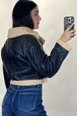 Campera tipo aviador de cuero sintético marrón con interior y detalles de corderito beige. Presenta cierre frontal cruzado, cuello solapa amplio, puños con corderito y cinturón ajustable en la cintura con hebilla metálica.