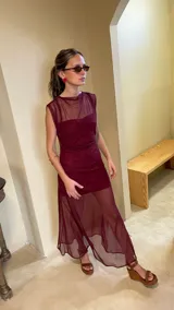 Vestido largo de microtul bordó, sin mangas, con escote alto tipo buche y diseño transparente con forro corto interno que genera un efecto translúcido en la falda.