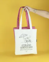 Bolso de mano tipo tote bag color crudo, con asas largas y estampado con motivos veraniegos en azul: sombrilla, reposera, perro caniche y texto "Verano Uruguayo".