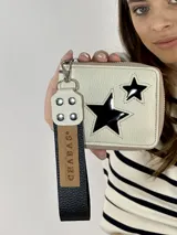 Billetera de cuero color crema con cierre perimetral y tira de cuero marrón con mosquetón y remaches. Presenta dos estrellas caladas en el frente, una grande y una pequeña.