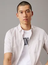 Camisa de manga corta Calvin Klein confeccionada en popelín de algodón elástico con diseño de rayas verticales en tonos beige y blanco. Presenta cuello en punta, cierre frontal de botones, bolsillo de parche en el pecho con logo bordado y bajo recto.