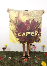 Pañuelo cuadrado de gran tamaño con estampado floral artístico en tonos amarillos y bordó, que incluye la palabra 'CAPER' en el centro y en los bordes.