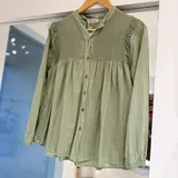 Camisa verde de manga larga con diseño amplio y fluido, cuello mao y cierre frontal con botones. Presenta detalles de nido de abeja en el canesú y fruncidos en los hombros.