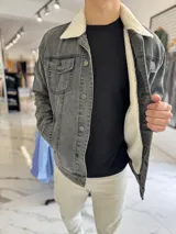 Chaqueta de jean color gris con interior y cuello de corderito sintético blanco, cierre frontal con botones metálicos y bolsillos en el pecho.