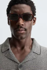 Camisa de punto de manga corta con cuello bowling y cierre frontal de botonadura. Presenta un patrón de rayas horizontales finas en blanco y negro, con ribetes negros en cuello, puños y bajo.