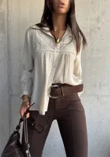 Camisa de color beige con cuello clásico, cierre frontal de botones y detalle de bordado geométrico en el pecho. Presenta mangas largas con puños y un diseño de tela con textura ligera.
