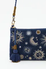 Cartera tipo sobre de algodón azul marino con bordados de motivos celestiales como lunas y estrellas en tonos dorados y plateados. Incluye una correa larga trenzada, cierre superior con cremallera y un detalle de borla decorativa en el lateral.