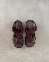 Sandalias de cuero color marrón con suela de goma negra, tiras anchas y hebilla metálica.