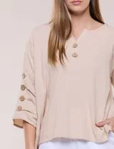 Blusa Erika de algodón con estampado de rayas en tonos azules, rosados y amarillos. Presenta cuello redondo con escote en V y botones frontales, mangas cortas decoradas con una fila de botones en los laterales y dobladillo asimétrico.