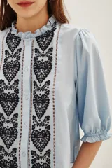 Camisa de algodón color celeste con cuello alto con volados, mangas tres cuartos abullonadas y cierre frontal con botones. Presenta un diseño con dos franjas verticales de bordado intrincado en color negro en el frente.