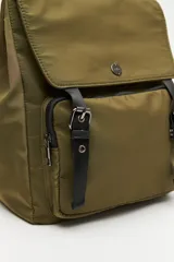Mochila urbana de nylon color beige, con solapa superior, bolsillo frontal con cierre y correas decorativas con hebillas metálicas. Incluye manija superior y correas ajustables.
