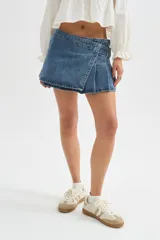 Minifalda de jean negra con diseño cruzado, cierre de hebilla metálica y detalle de tablones plisados en el lateral.