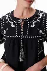 Blusa negra de algodón con bordados blancos a contratono, cuello redondo con lazo y borlas decorativas, y mangas cortas.
