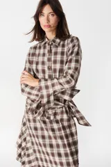 Camisa de corte holgado con estampado de cuadros en tonos marrones y beige. Presenta cuello clásico, cierre frontal con botones y mangas largas con aberturas laterales que permiten anudar la prenda en la cintura.