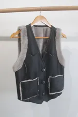 Chaleco de cuero genuino color negro con interior y detalles de piel sintética en color gris. Presenta cierre frontal con botones, cuello en V y dos bolsillos delanteros con borde de piel.