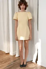 Vestido corto color amarillo claro, con cuello barco, mangas cortas abullonadas y pliegues en la parte delantera. Presenta falsos bolsillos de vivo y costuras marcadas. Cierre en la espalda con cremallera oculta.