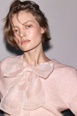 Saco de punto suave color rosado, de manga corta y cuello redondo. Presenta cierre frontal con botones forrados y una moña decorativa de organza transparente en la parte superior. Posee terminaciones acanaladas en mangas y ruedo.