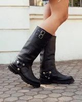 Bota de caña alta confeccionada en cuero negro, con suela track de goma. Presenta un diseño versátil con dos hebillas ajustables en la parte superior de la caña y una correa decorativa con tachas metálicas y argollas en la zona del tobillo.