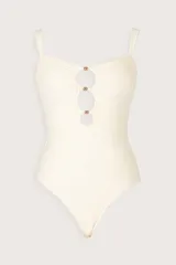 Traje de baño enterizo color crema con diseño bandeau, fruncidos laterales y tres aberturas circulares frontales decoradas con detalles metálicos brillantes.