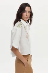 Camisa crop blanca de lino con silueta relajada, cuello clásico y mangas cortas. Presenta bordados de tigres en el frente y detalles ornamentales en los hombros.