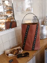 Bolso tipo matera bucket de cuero color marrón, con diseño cilíndrico y dos franjas verticales de cuero negro con tachas metálicas plateadas. Cuenta con un asa superior fija y un compartimento interior divisor.