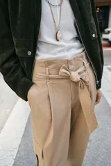 Pantalón de tiro alto estilo paperbag en color beige, con pinzas frontales y cinturón de la misma tela. Presenta bolsillos laterales tipo cargo con solapa y pernera ajustable con botones en el bajo.