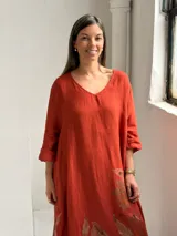 Vestido largo color terracota con escote en V, mangas tres cuartos y diseño amplio, decorado con un estampado floral grande en tono marrón en la parte inferior.