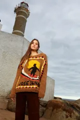 Sweater tejido de lana color marrón con cuello redondo y puños ajustados. Presenta un diseño con un sol amarillo, un surfista negro y la inscripción "José Ignacio" en el frente.