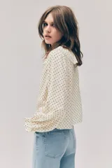 Blusa de viscosa color beige con estampado de lunares negros. Presenta cuello con volados, cordón fino para ajustar, botonadura frontal y mangas largas con puños elásticos fruncidos.