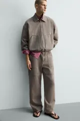 Pantalón de corte relajado en color marrón con efecto lavado. Presenta bolsillos frontales tipo cargo, cierre con cremallera y botón, y una línea horizontal de tachas metálicas a tono en la parte superior de las piernas.