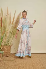 Vestido largo estampado con motivos florales en tonos pastel y detalles de cadenas doradas. Presenta cuello mao, cierre frontal con botones variados, mangas largas con puños elásticos y un cinturón a tono con hebilla dorada que realza la cintura. El ruedo del vestido tiene una franja de color celeste.