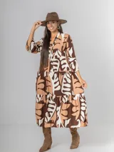 Vestido largo de manga tres cuartos, con cuello camisero y botones frontales. Presenta un estampado abstracto en tonos marrón oscuro, beige y blanco. El calce es amplio y tiene un ruedo con volado.