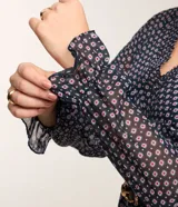 Vestido corto azul marino con estampado de flores blancas y rojas, escote en V, mangas largas con puños elásticos y falda con volantes. Incluye cinturón negro con hebilla dorada.