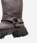 Bota marrón de cuero sintético, estilo biker, con caña media, puntera redonda, suela track y detalle de arnés con hebilla metálica.