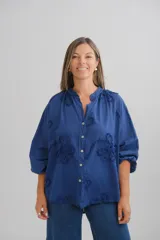 Blusa azul marino de corte holgado, con cuello mao y cierre frontal de botones. Presenta mangas tres cuartos con puños elásticos y un delicado diseño de bordados florales en relieve distribuidos por toda la prenda.