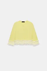 Chaqueta de punto color amarillo claro, con cuello redondo y mangas por debajo del codo. Presenta detalles de acabado en tejido crochet blanco en el ruedo y los puños, con cierre frontal de botones forrados al tono.