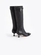 Bota de caña alta confeccionada en cuero vacuno liso, con taco laqueado de altura intermedia y punta fina. Presenta detalles de hebillas metálicas en la caña y en la puntera, incluyendo un dije en forma de corazón.