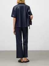 Camisa de manga corta confeccionada en cuero sintético color azul marino. Presenta un diseño de línea recta, cuello solapa, cierre frontal con botones y un bolsillo aplicado en el pecho.