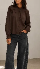 Camisa marrón oscuro de manga larga, con detalle de volados en cuello alto, a lo largo de la botonadura frontal y en los puños elásticos. Presenta frunces en el canesú delantero y cierre frontal con botones.